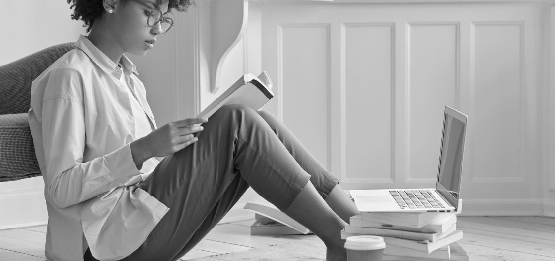 Sideways shot of hard working black entrepreneur wants to be promoted, studies business literature, sits on floor with papers in front of laptop computer, being sole proprietor of prosperous company